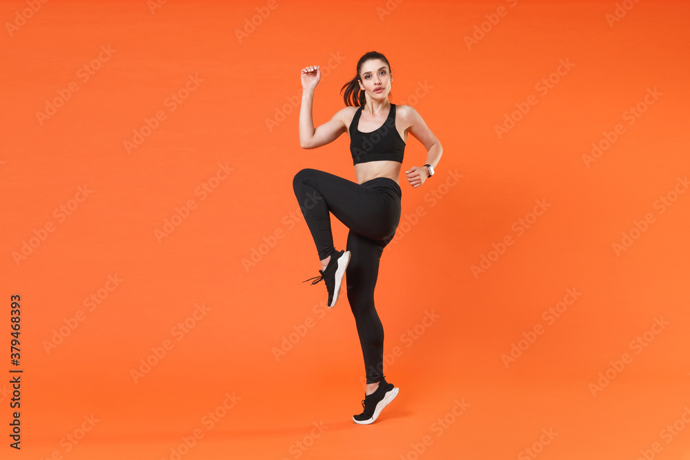 Full length side view portrait beautiful young fitness sporty woman in black sportswear posing training jumping dancing doing cardio exercising rising hands legs isolated on orange background studio.