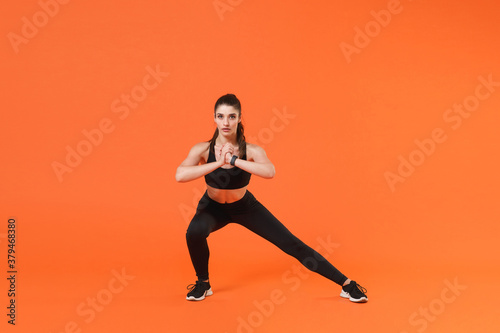 Full length portrait of beautiful young fitness sporty woman in black sportswear posing training working out doing stretching exercise lunge looking camera isolated on orange color background studio.