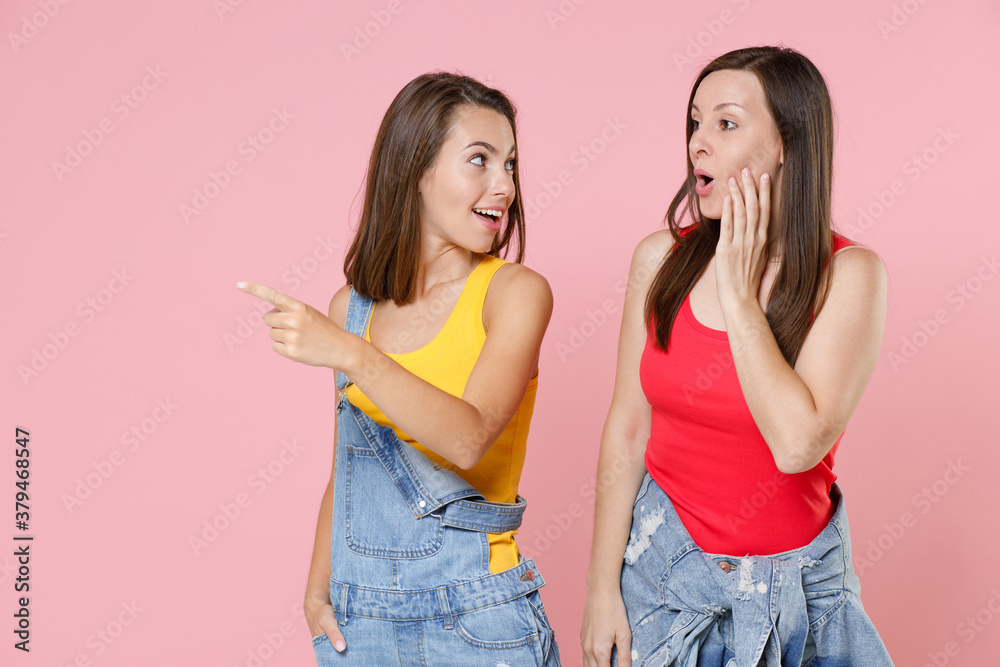 Two shocked amazed young women friends 20s in casual denim clothes stand pointing index finger aside on mock up copy space put hand on cheek isolated on pastel pink colour background, studio portrait. Two shocked amazed young women friends 20s in casual denim clothes stand pointing index finger aside on mock up copy space put hand on cheek isolated on pastel pink colour background, studio portrait.
