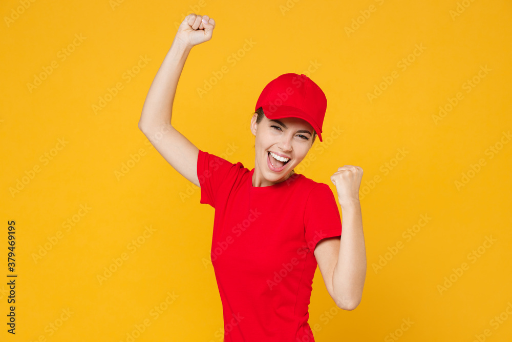 Fun delivery employee woman in red cap blank t-shirt uniform workwear work courier in service during quarantine coronavirus covid-19, doing winner gesture isolated on yellow background studio portrait