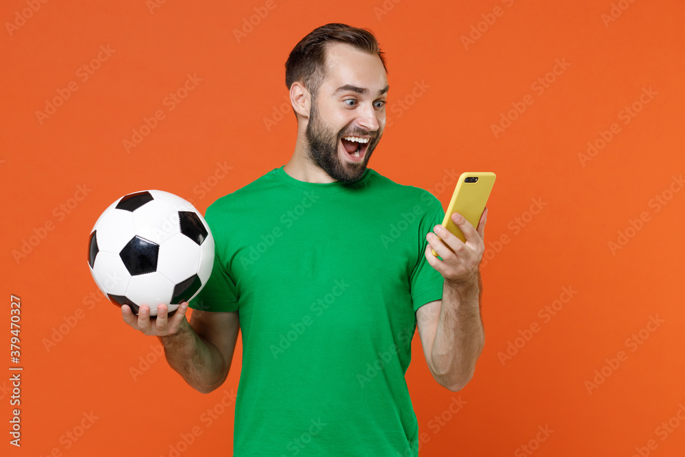 Φωτογραφία Surprised young man football fan in green t-shirt cheer up support favorite team with soccer ball using mobile cell phone isolated on orange background studio