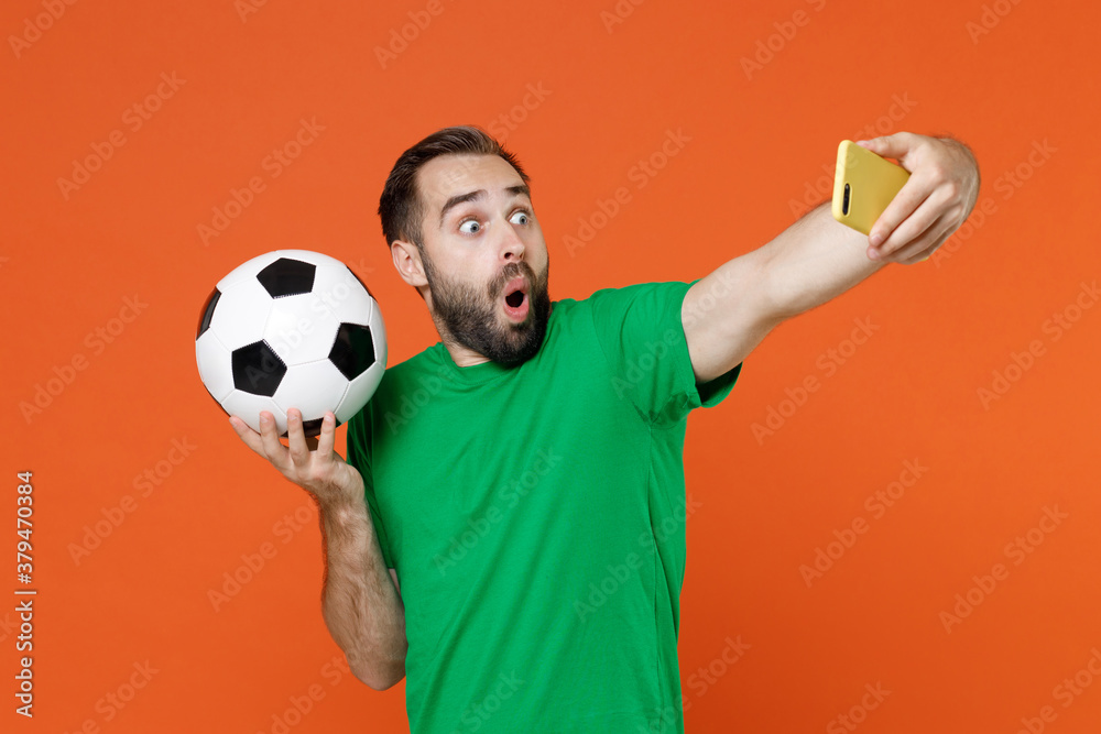 Φωτογραφία Shocked young man football fan in green t-shirt cheer up support favorite team with soccer ball doing selfie shot on mobile phone isolated on orange background