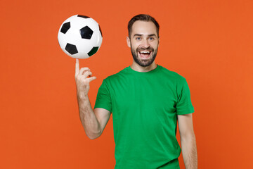 Funny young man football fan in green t-shirt cheer up support favorite team hold rotating soccer ball on finger isolated on orange background studio portrait. People sport leisure lifestyle concept.