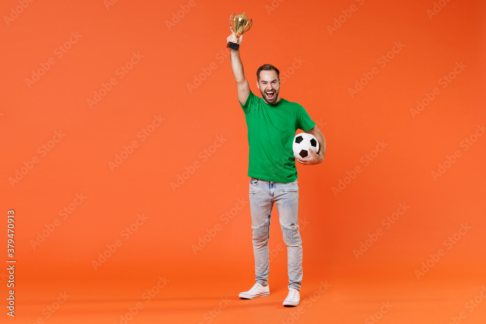 Full length portrait happy young man football fan in green t-shirt cheer up support favorite team with soccer ball hold golden cup isolated on orange background studio. People sport leisure concept. Full length portrait happy young man football fan in green t-shirt cheer up support favorite team with soccer ball hold golden cup isolated on orange background studio. People sport leisure concept.