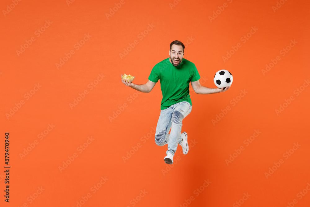 Full length portrait screaming man football fan in green t-shirt cheer up support favorite team with soccer ball hold bowl of chips jumping isolated on orange background. People sport leisure concept.