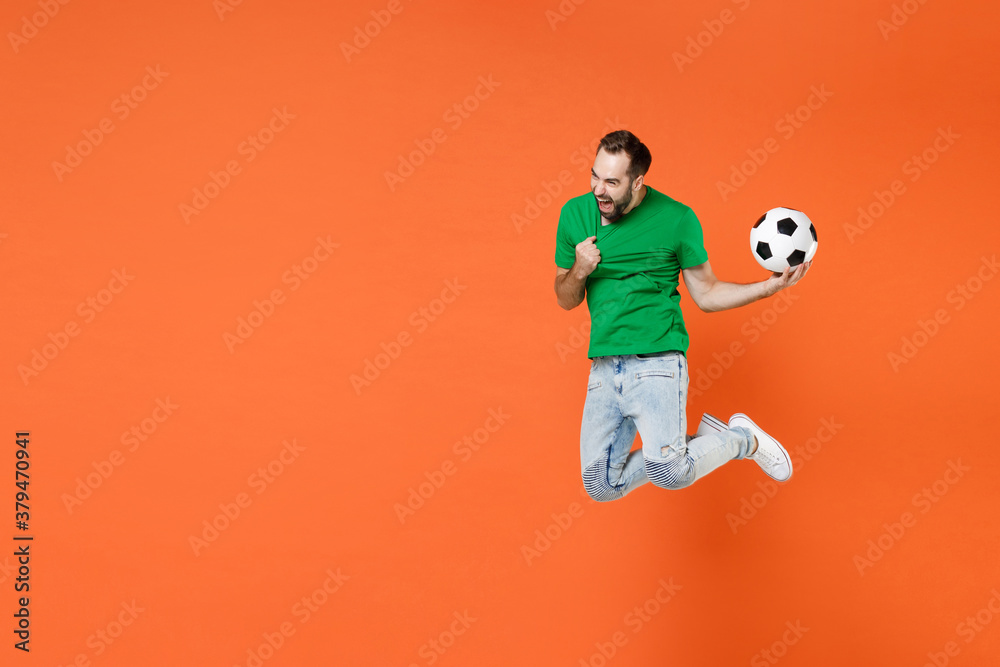 Full length portrait crazy man football fan tearing green t-shirt cheer up support favorite team with soccer ball jumping screaming isolated on orange background studio. People sport leisure concept.