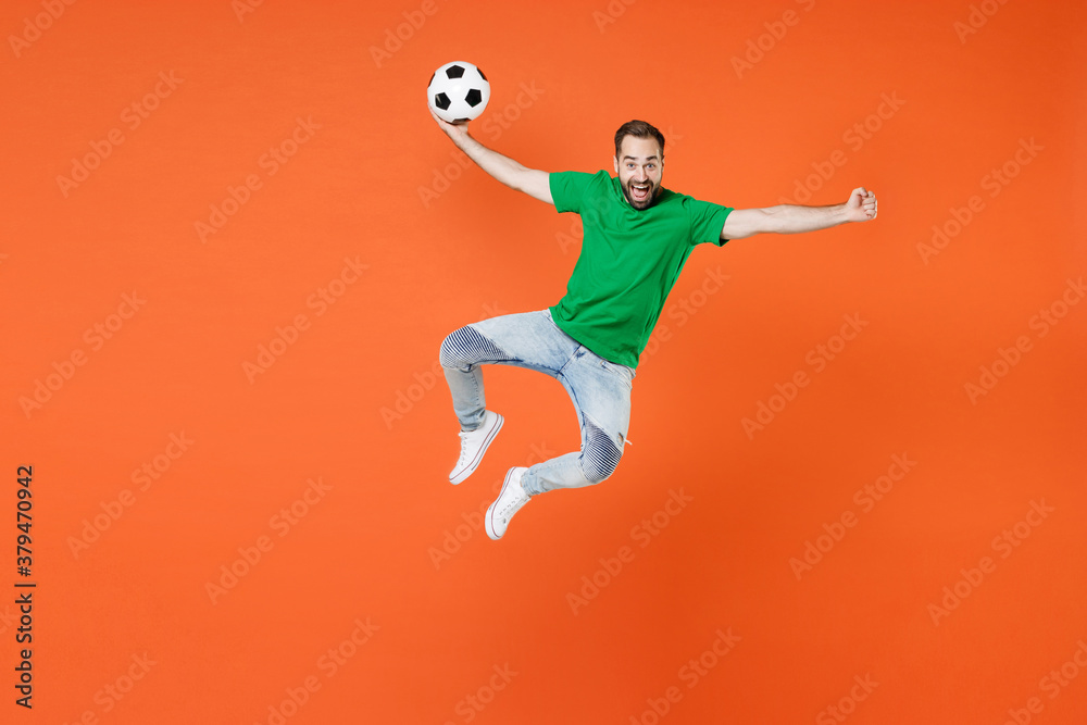Full length portrait of excited man football fan in green t-shirt cheer up support favorite team with soccer ball jumping clenching fist isolated on orange background. People sport leisure concept.