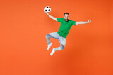 Full length portrait of excited man football fan in green t-shirt cheer up support favorite team with soccer ball jumping clenching fist isolated on orange background. People sport leisure concept.