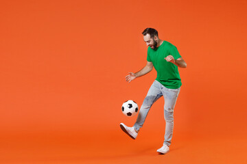 Full length portrait funny young man football fan in basic green t-shirt cheer up support favorite team kicking juggling soccer ball isolated on orange background studio. People sport leisure concept.