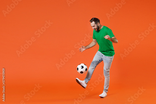 Fototapeta Naklejka Na Ścianę i Meble -  Full length portrait funny young man football fan in basic green t-shirt cheer up support favorite team kicking juggling soccer ball isolated on orange background studio. People sport leisure concept.