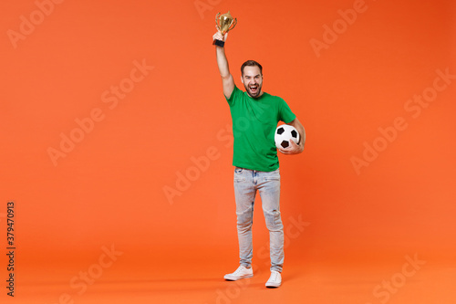 Fototapeta Naklejka Na Ścianę i Meble -  Full length portrait happy young man football fan in green t-shirt cheer up support favorite team with soccer ball hold golden cup isolated on orange background studio. People sport leisure concept.