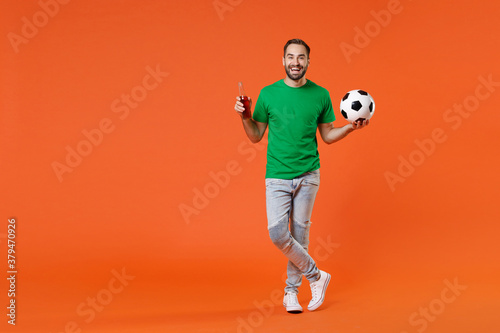 Fototapeta Naklejka Na Ścianę i Meble -  Full length portrait funny young man football fan in green t-shirt cheer up support favorite team with soccer ball hold beer bottle isolated on orange background studio. People sport leisure concept.