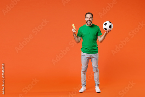 Fototapeta Naklejka Na Ścianę i Meble -  Full length portrait of cheerful man football fan in basic green t-shirt cheer up support favorite team with soccer ball using mobile phone isolated on orange background. People sport leisure concept.