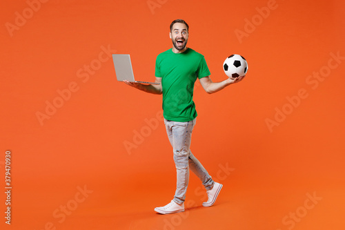 Fototapeta Naklejka Na Ścianę i Meble -  Full length portrait excited man football fan in basic green t-shirt cheer up support favorite team with soccer ball using laptop computer isolated on orange background. People sport leisure concept.