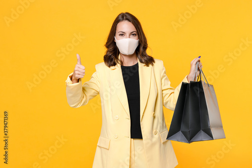 Young woman in suit jacket face mask to safe from coronavirus virus covid-19 hold package bags with purchases after shopping showing thumb up isolated on yellow background studio. Black friday sale.