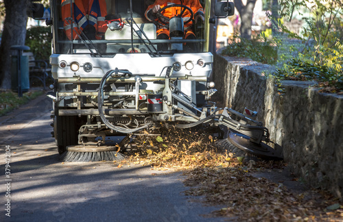 Fototapeta Naklejka Na Ścianę i Meble -  New generation of small electric street sweeper removing fallen leaves in body at autumn city park. Municipal urban services using ecology green vehicle lorry to clean streets from foliage.