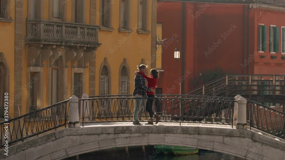 Handsome loving man kissing and hugging his charming asian girlfriend standing on the ancient Venetian bridge during their honeymoon. Travelling to Europe as the best way of spending the vacation.