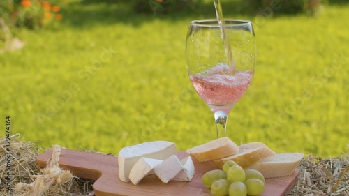 Close up rose wine pouring into a glass from a bottle. Cheese, bread and grape on wooden cutting board. Summer day in the garden. Macro shot with slide or move camera
