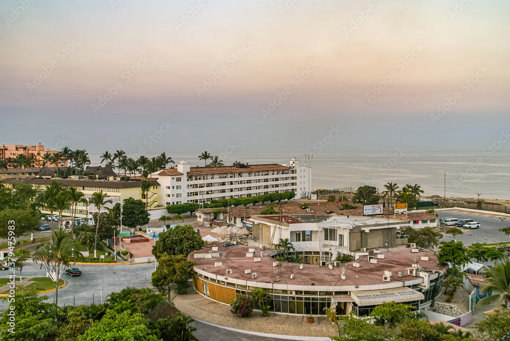 Puerto Vallarta, Mexico - April 25, 2008: Morning yellow-orange light over ocean with circular port authority offices up front on harbor. Other buildings around with green foliage.