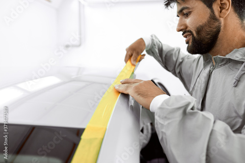 mechanic caucasian man with yellow paper tape plasters car for polishing it, paint and varnish, remove scratches in a vehicle wash and detailing workshop.