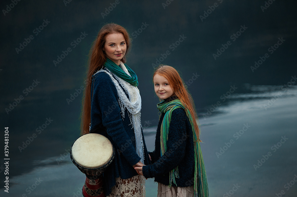 A young woman and a girl with red long hair walk along the lake in autumn.