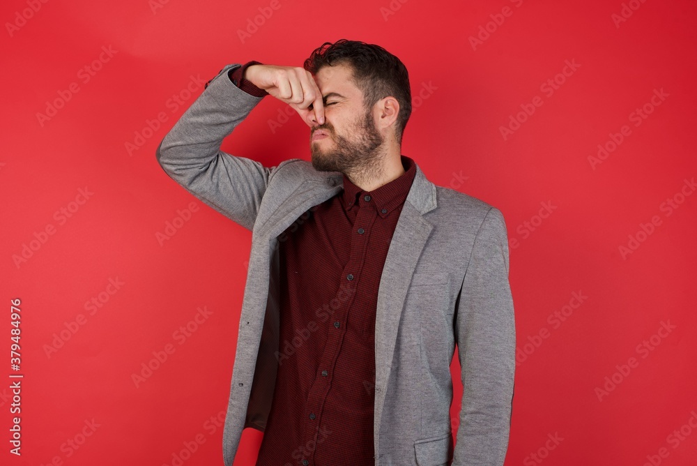 Young caucasian businessman wearing casual clothes standing over isolated red background smelling something stinky and disgusting, intolerable smell, holding breath with fingers on nose. Bad smell Young caucasian businessman wearing casual clothes standing over isolated red background smelling something stinky and disgusting, intolerable smell, holding breath with fingers on nose. Bad smell