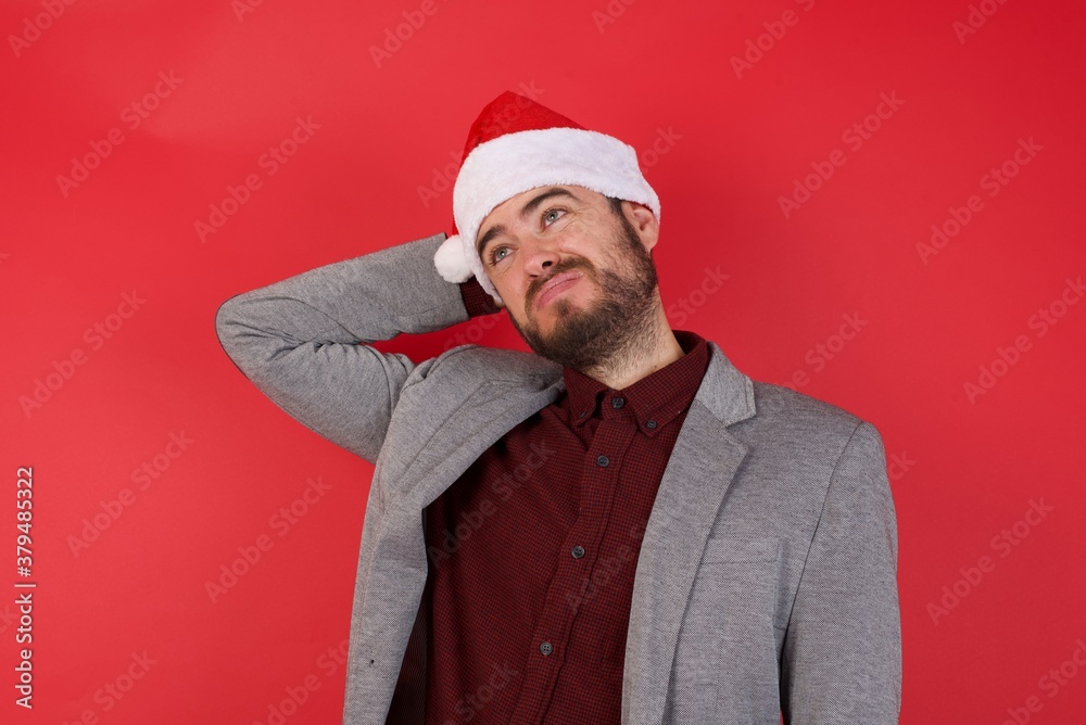 Young caucasian businessman wearing casual clothes and Santa hat standing over isolated red wall confuse and wonder about question. Uncertain with doubt, thinking with hand on head. Pensive concept. Young caucasian businessman wearing casual clothes and Santa hat standing over isolated red wall confuse and wonder about question. Uncertain with doubt, thinking with hand on head. Pensive concept.