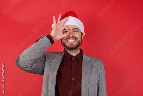 Young caucasian businessman wearing casual clothes and Santa hat standing over isolated red wall doing ok gesture with hand smiling, eye looking through fingers with happy face.