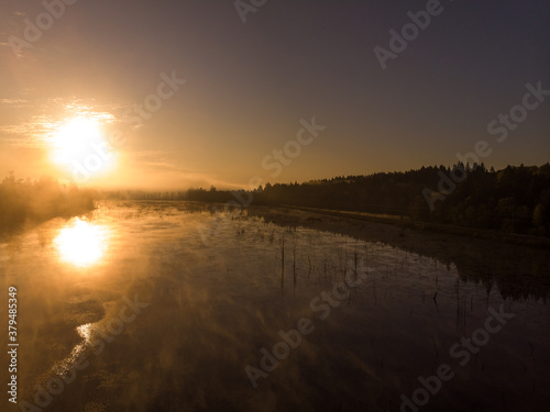 Amazing sunrise on the forest lake. The sun reflecting on the water. Early morning natural landscape. The lightly blue sky background.