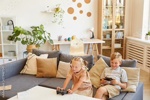 High angle portrait of cute girl with down syndrome playing video games with brother while sitting on couch in living room, copy space