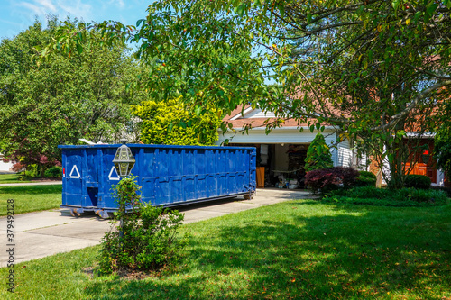An empty blue dumpster in the driveway of a house with its garage door open in a residential community photo