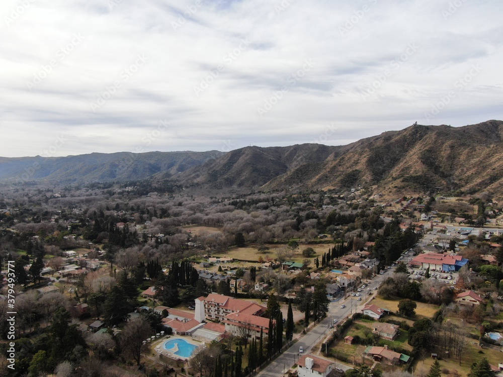 Vista aérea panorámica desde un dron, de un pueblo en un valle, con una cordillera cubierta de vegetación y nubes grises.
