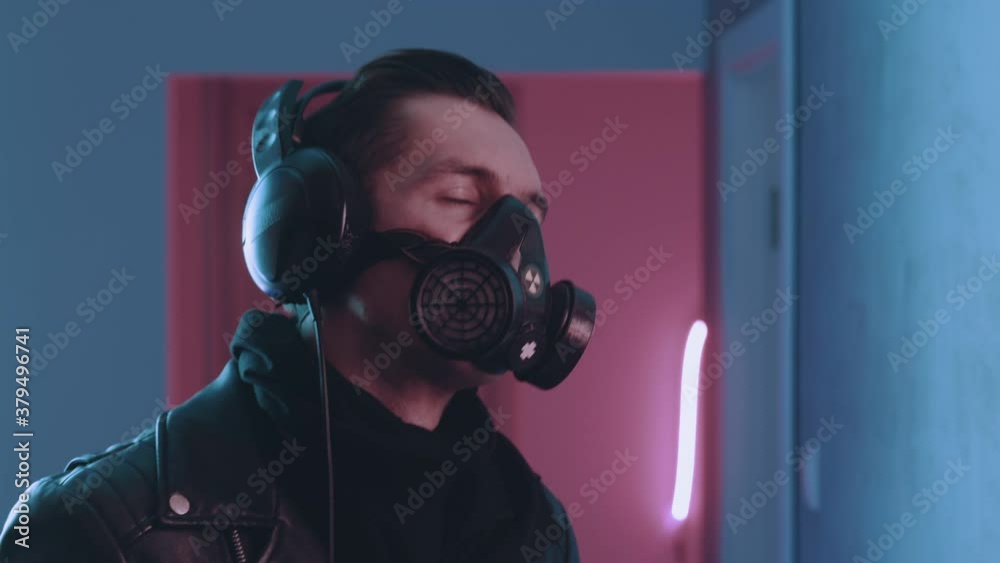 Cyberpunk portrait of brutal man in respirator enjoys listening to music with headphones in neon light. Young male in gas mask listening to music on blue and pink neon light background.
