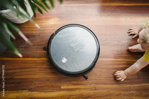 a robot vacuum cleaner cleaning the floor with a baby dressed in yellow and a plant