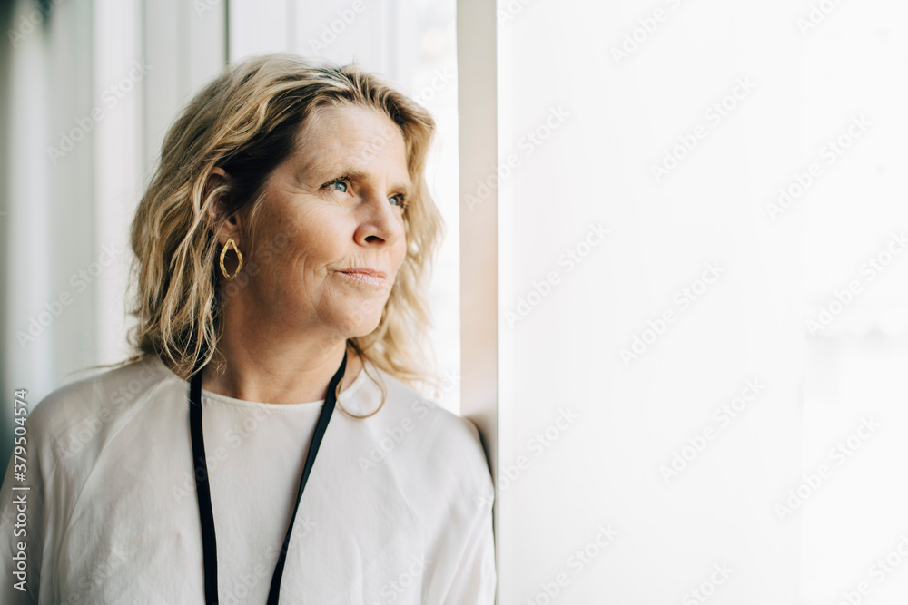 Mature entrepreneur looking away while leaning by window at workplace Mature entrepreneur looking away while leaning by window at workplace