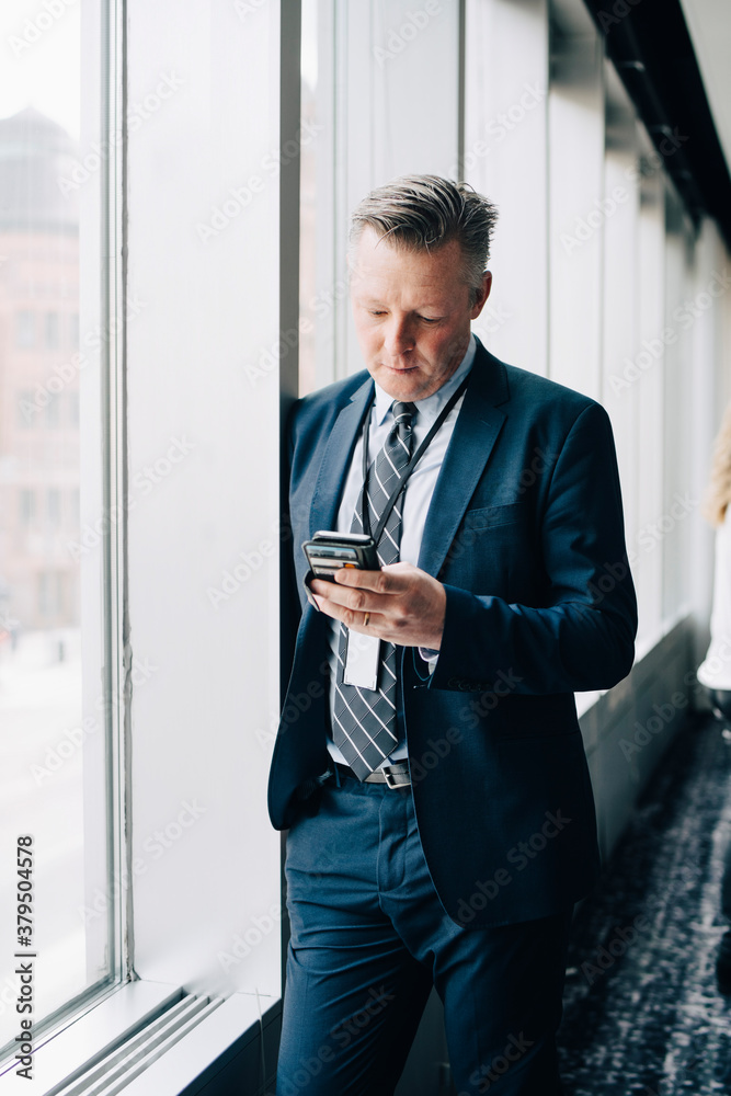Entrepreneur using phone while standing by window in office Entrepreneur using phone while standing by window in office
