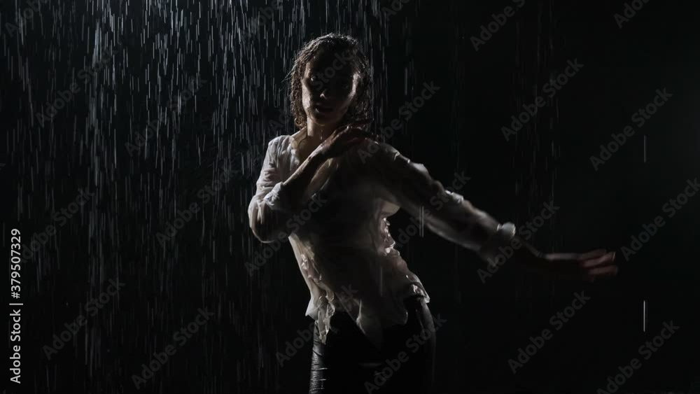 Beautiful young woman dancing in a white shirt and black short shorts dancing in the rain in the studio on a black background. Dancing on the water.