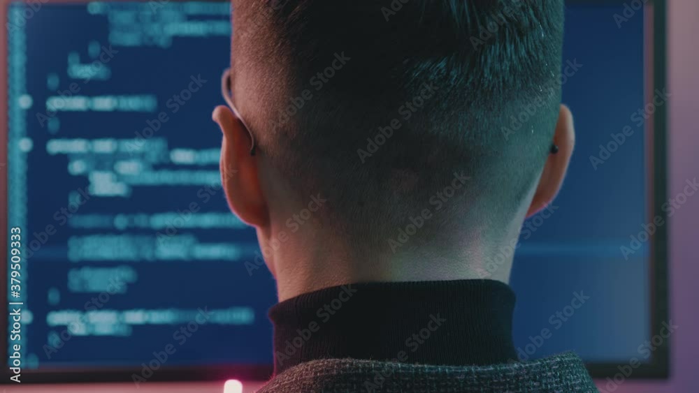 Rear view of man programmer coding network security software indoors in neon lights. Hacker typing code on computer screen in dark office at night