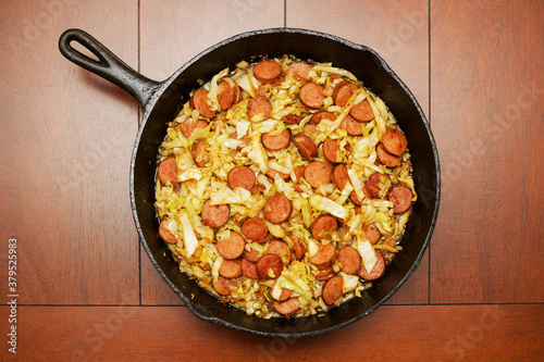 A cast iron skillet with fried cabbage and kielbasa sitting on a wood table. photo