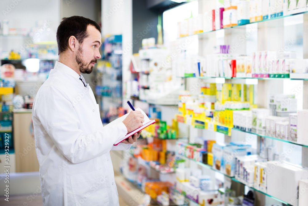 Young male pharmacist checking assortment of drugs in drugstore