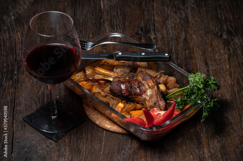 Roasted lamb leg with potatoes fresh cooked out of the oven. Delicious roasted ribs. Appetizing baked lamb shins from oven with vegetable garnish of french fries, parsley. Red wine. Dark wood table