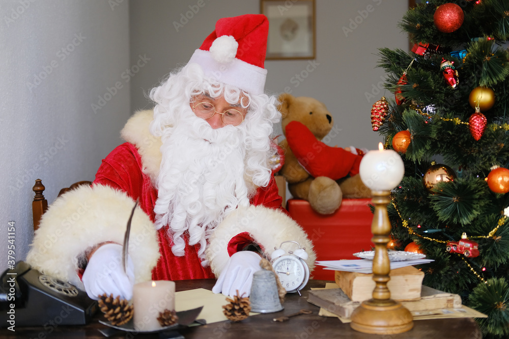 Fototapeta premium santa claus with a white beard in a red suit sits by a christmas tree decorated with balls, writes a letter with a pen, concept of christmas, waiting for gifts, celebrating
