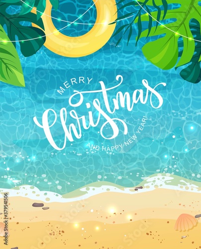 Merry Christmas hand lettering text for exotic New Year celebration. Summer seashore with sand and yellow swim ring, tropical leaves, top view. The wave rolls onto the sand, sea foam, blue water. 