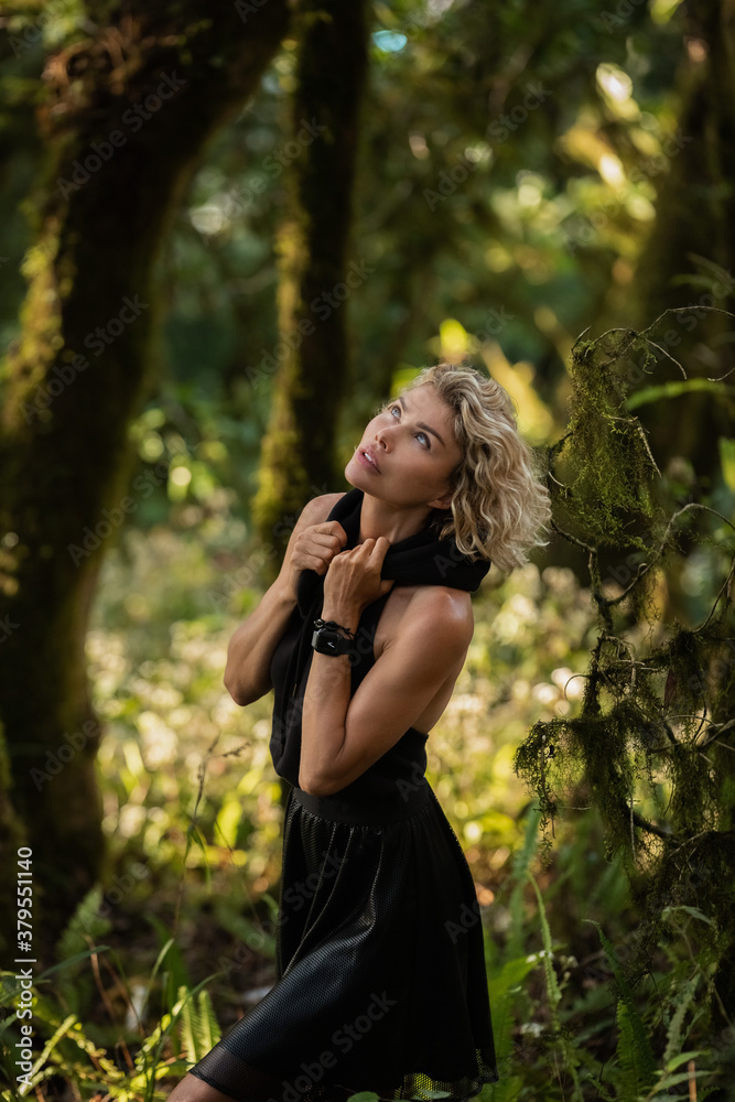  Brooding dreamy young girl blonde, black dress with a hood standing by a tree. The girl got lost in the woods. Trekking, fantasy, magical mystical forest. Girl standing by a tree. High quality photo