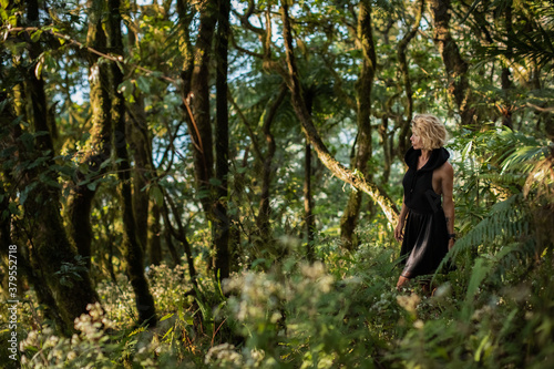  Brooding dreamy young girl blonde, black dress with a hood standing by a tree. The girl got lost in the woods. Trekking, fantasy, magical mystical forest. Girl standing by a tree. High quality photo
