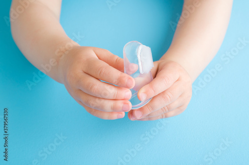 Baby hands holding transparent silicone fingertip toothbrush on light blue table background. Pastel color. Babies teeth hygiene. Closeup. Front view.