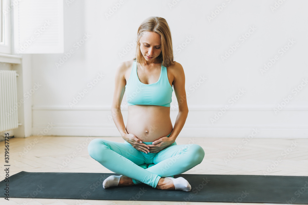 Happy young pregnant woman in sports uniform doing stretching in bright Studio