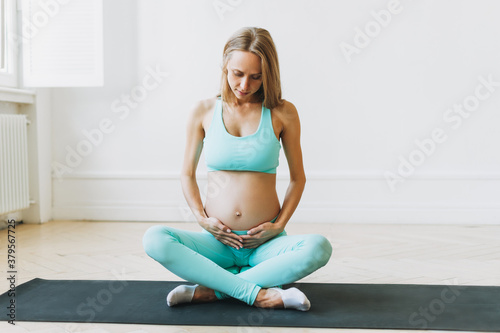 Happy young pregnant woman in sports uniform doing stretching in bright Studio