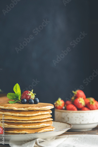 Pancake with srtawberry, blueberry and mint in ceramic dish, syrup from small ceramic jar and flowers on a dark wooden table photo