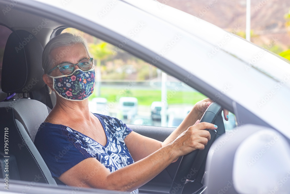 Naklejka premium Senior woman wearing a flowered surgical mask because of the covid19 coronavirus ready to drive her gray car looking at camera - concept of active retirees